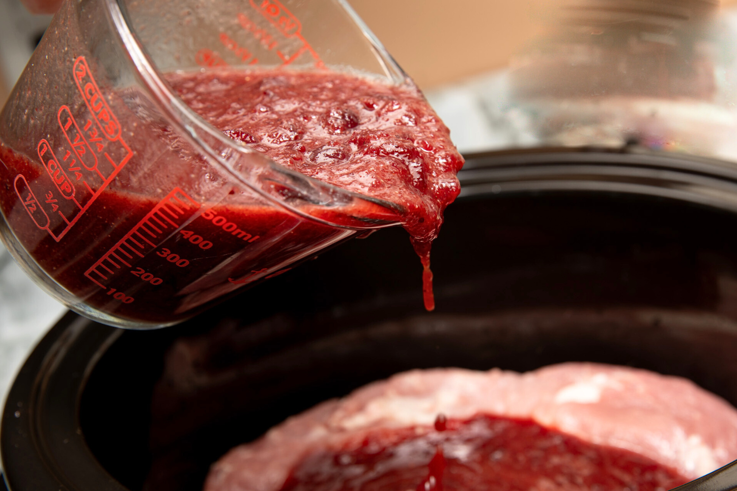Glass measuring cup pouring cranberry sauce mixture on top of pork tenderloin.