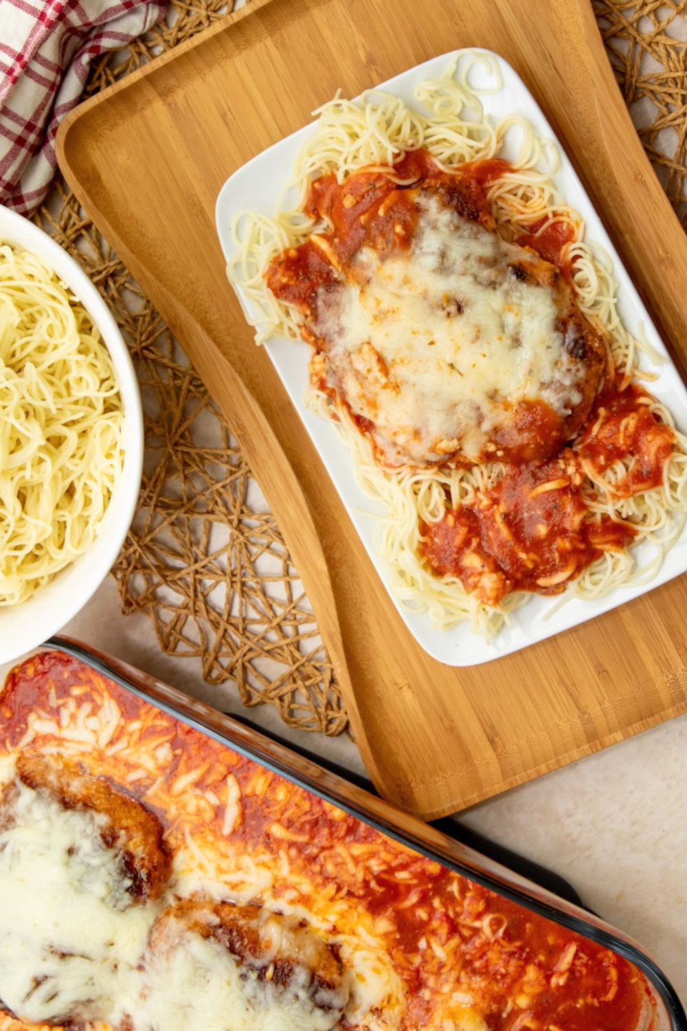 Wooden platter with a plate of pork cube steak parmesan and pasta surrounded by red sauce, next to glass serving dish.