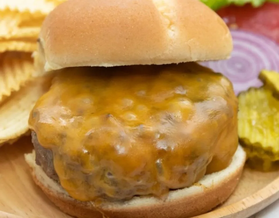A cheesy pork and apple burger on a cutting board with toppings and chips.