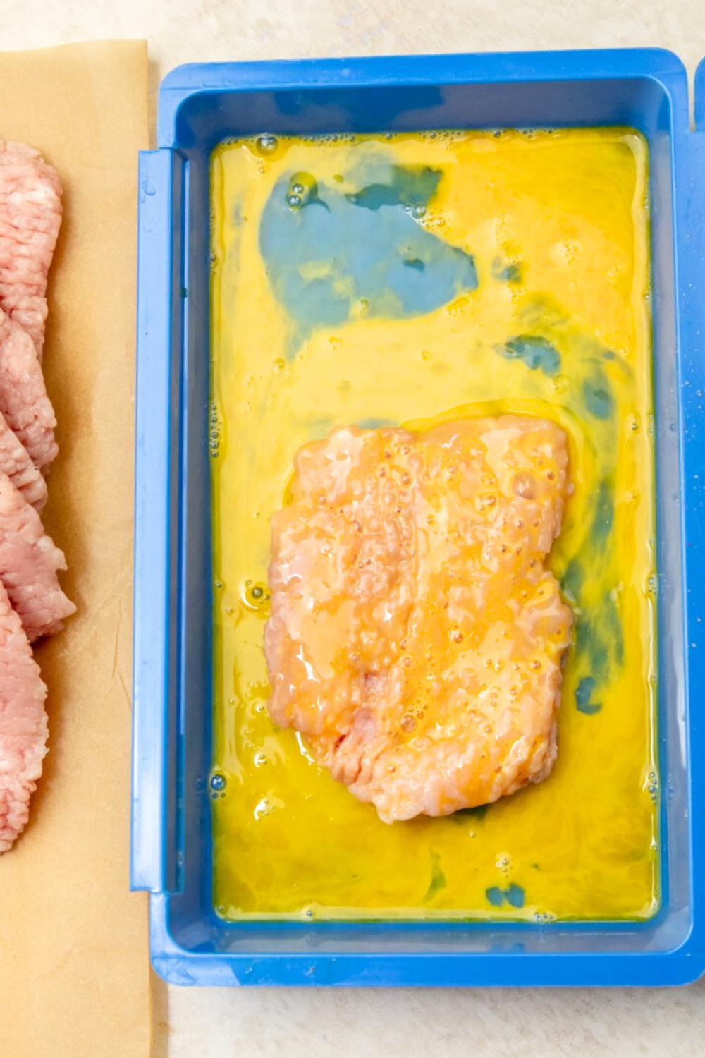 Pork cube steak surrounded by egg wash in blue coating tray.