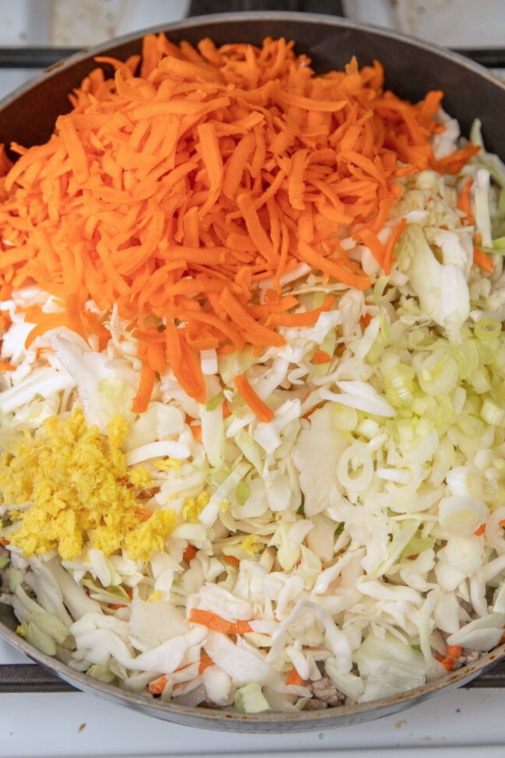 coleslaw-carrots-frying Shredded cabbage, carrots, onion and ginger on top of ground pork in skillet.