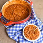 Blue and white table cloth with pot of pork and beans and white bowl of beans.